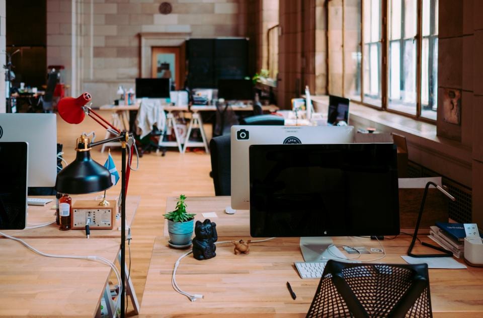 Open space lumineux aux grands bureaux partagés en bois clair, avec écrans, lampes d’architecte et petites plantes