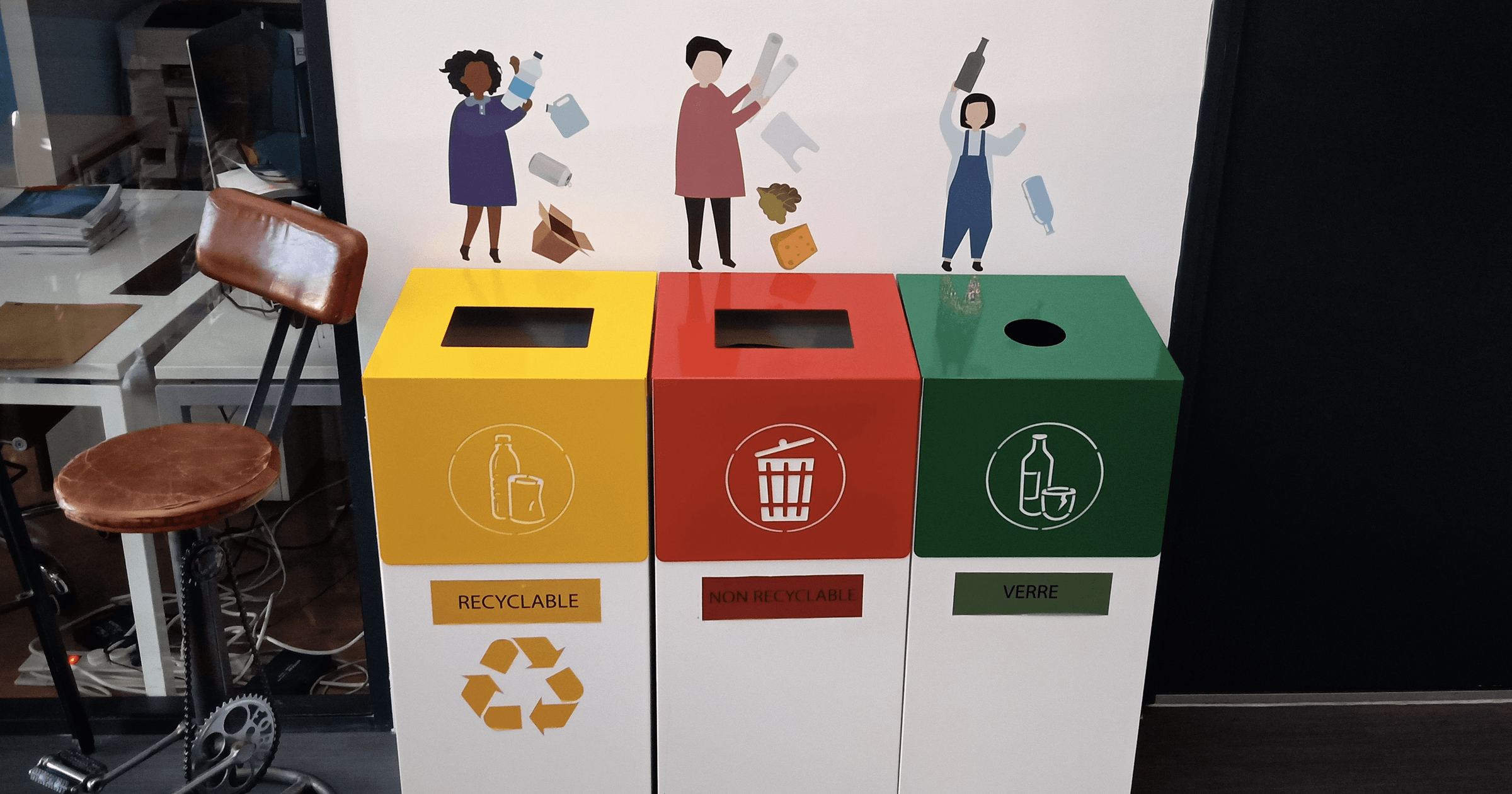 Trois poubelles de tri Kollori côte à côte : jaune pour les recyclables, rouge pour le non recyclable, verte pour le verre, sous des illustrations murales