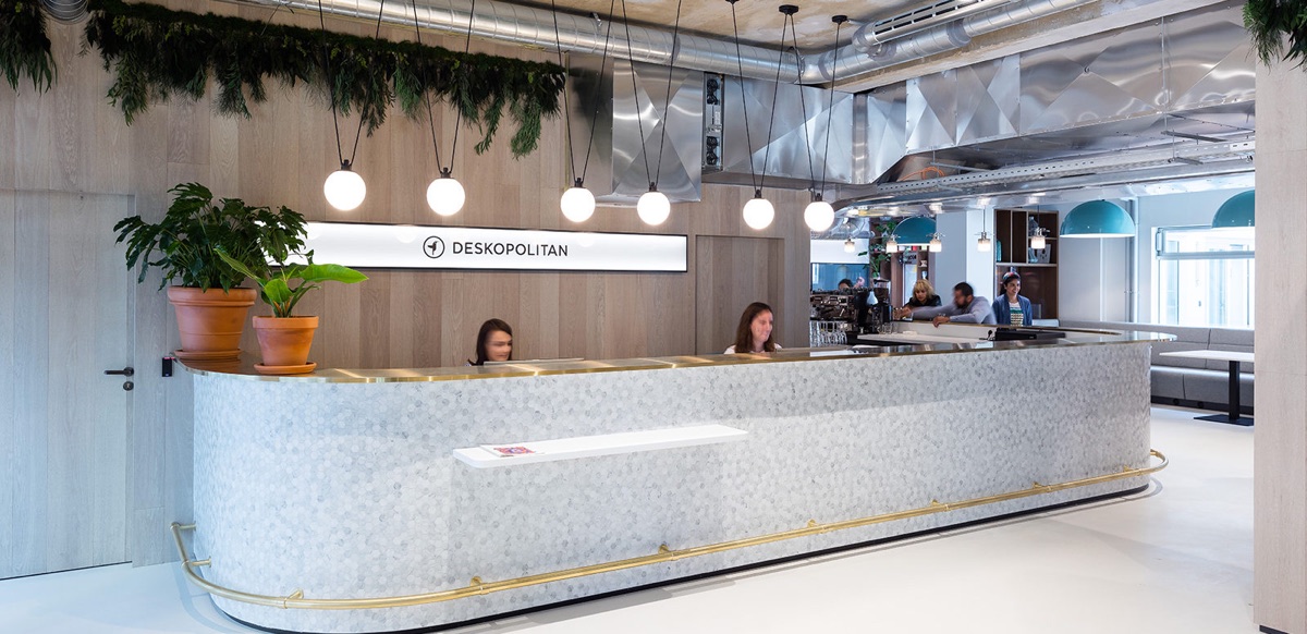 Accueil de Deskopolitan Voltaire : long comptoir arrondi en mosaïque de marbre blanc bordé de laiton, suspensions à globes, plantes vertes et mur habillé de bois clair, avec le bar à café et un espace de travail en arrière-plan sous des gaines techniques apparentes