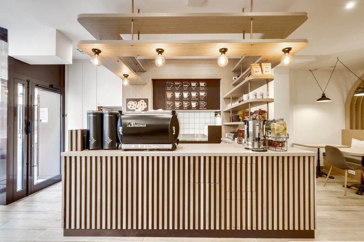Salle du café-coworking Hubsy République : banquettes grises garnies de coussins blancs le long de grandes tables en bois clair, sous une structure de poutres en bois en forme de toit d’où pendent des plantes grimpantes et une ampoule rouge