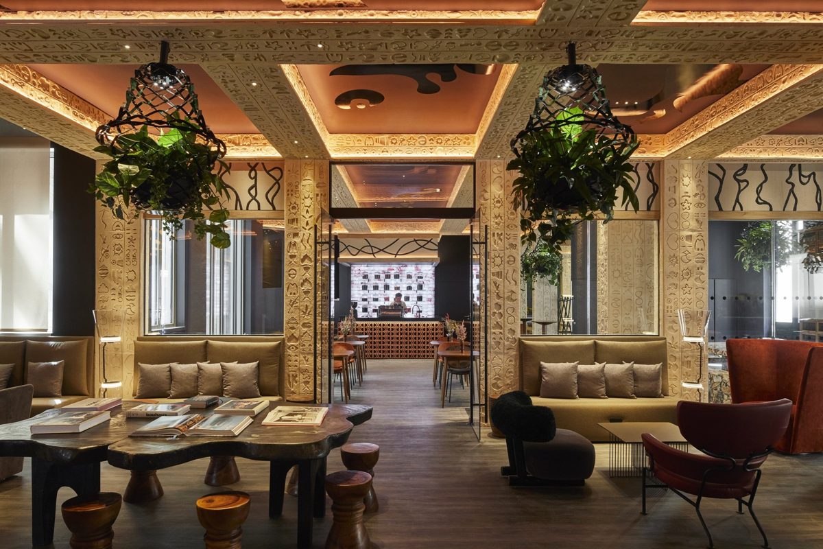Lobby de Kwerk Madeleine : banquettes beiges à coussins, table en bois brut couverte de livres, tabourets en bois, piliers et caissons sculptés de motifs gravés, suspensions végétales, avec un comptoir en perspective