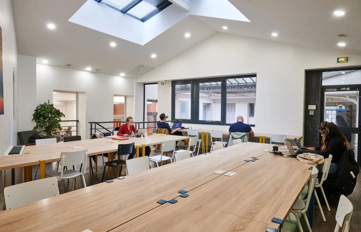 Grande salle de travail de La Permanence Alésia, à Paris 14e : une verrière au plafond, de longues tables en bois avec des chaises blanches et des coworkers sur leur ordinateur portable