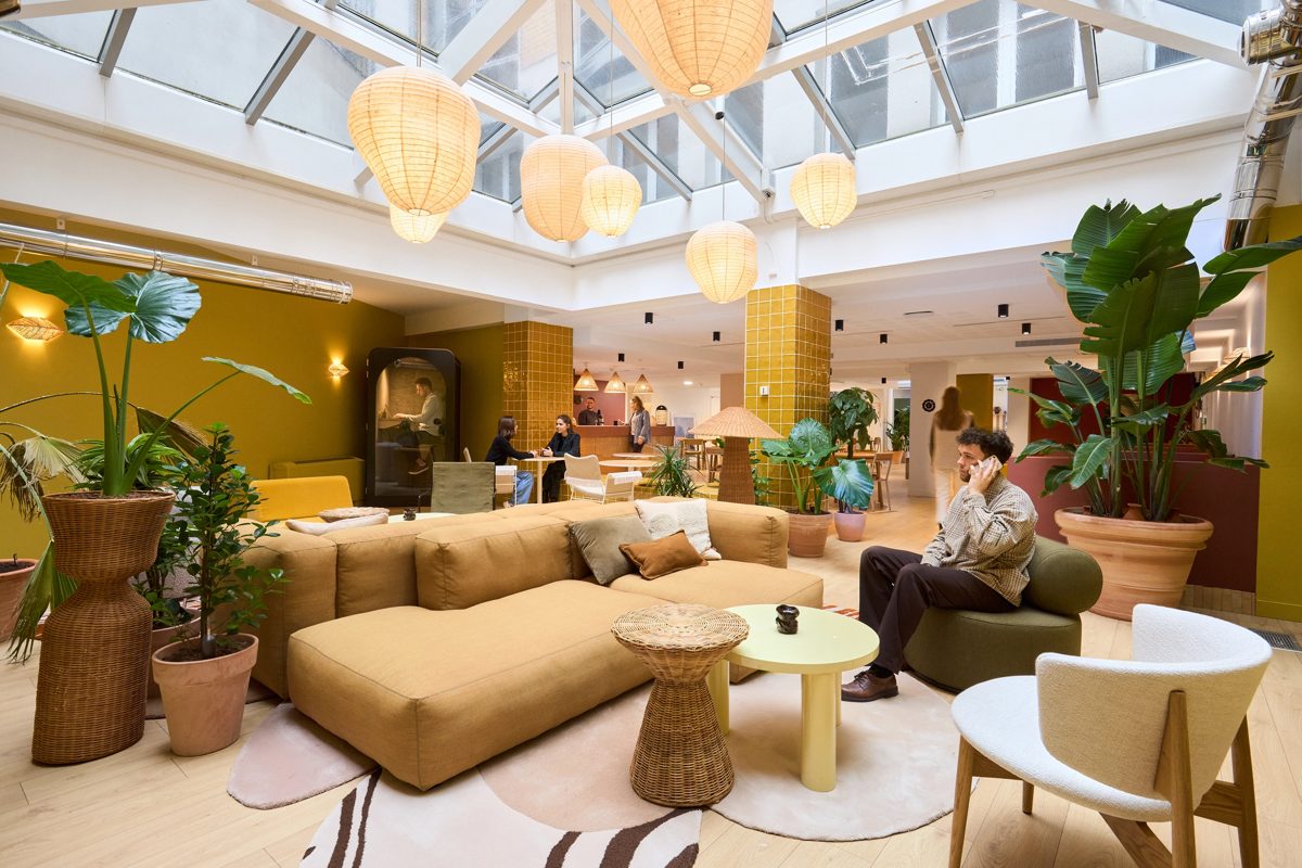 Salon de Morning Bourse sous une verrière : grand canapé moutarde, suspensions en papier, plantes vertes en pots, colonnes carrelées jaunes et comptoir café au fond