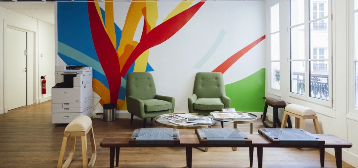 Espace détente du premier étage de Mozaik Coworking, rue de la Chaussée d’Antin à Paris 9e : deux fauteuils verts, des tables basses, des bancs et des tabourets en bois devant une grande fresque colorée, à côté de hautes fenêtres