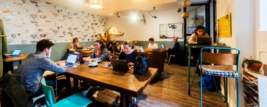 L’open space du Nuage Café, rue des Carmes (Paris 5e) : des coworkers sur leur ordinateur portable autour de grandes tables en bois, devant un papier peint à motifs de montgolfières, avec une table haute et un échiquier au fond