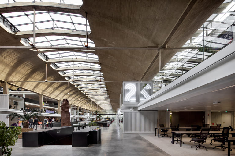Intérieur de Station F à Paris : la grande nef aux voûtes de béton percées de verrières, l’allée centrale et des postes de travail avec fauteuils noirs sous une mezzanine vitrée