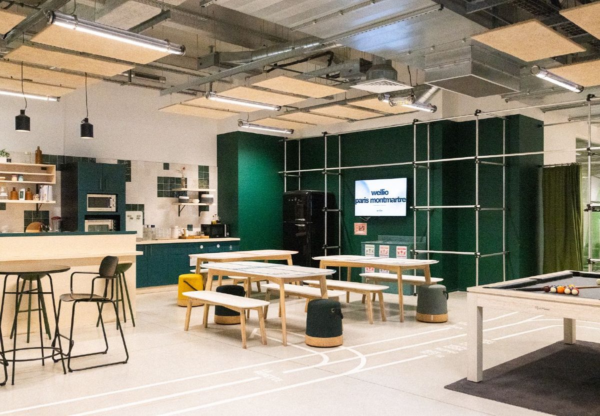 Espace commun de Wellio Paris Montmartre : cuisine ouverte, tables en bois et bancs, poufs colorés, billard, murs vert sapin garnis d’un filet de tubes métalliques et lignes de terrain de sport peintes au sol