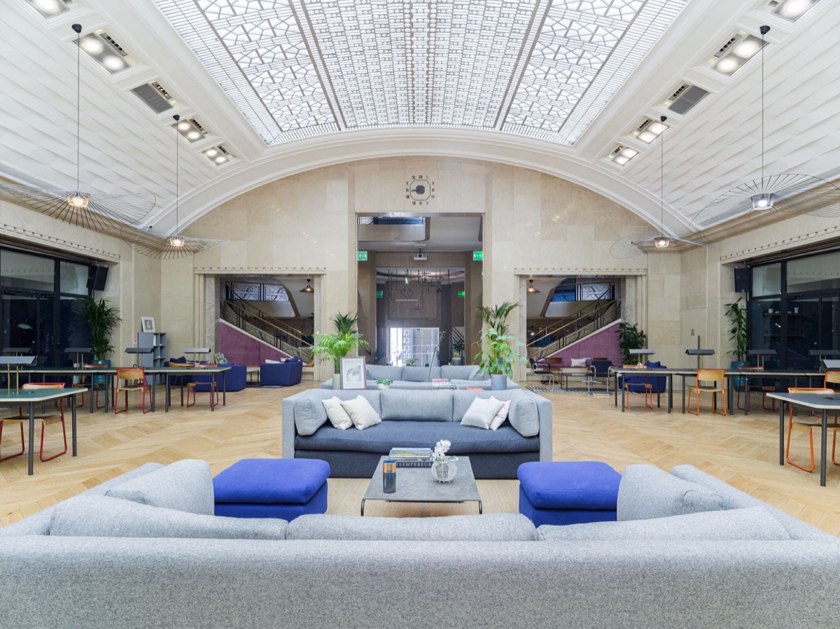 L’atrium du WeWork du 33 rue La Fayette sous sa grande verrière voûtée Art déco : canapés gris et poufs bleus au centre, tables de travail et chaises orange sur un parquet à chevrons, et deux escaliers au fond