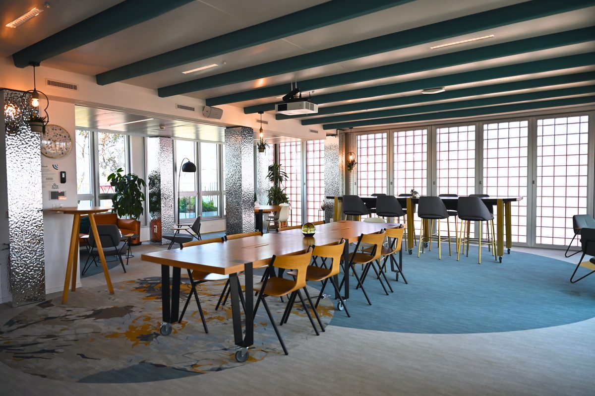 Espace de travail du Wojo Paris 13e - BnF : tables hautes en bois le long de grandes baies vitrées, banquette verte bordée de plantes, sol en terrazzo coloré et plafond peint en ciel nuageux, où quelques personnes travaillent sur ordinateur portable