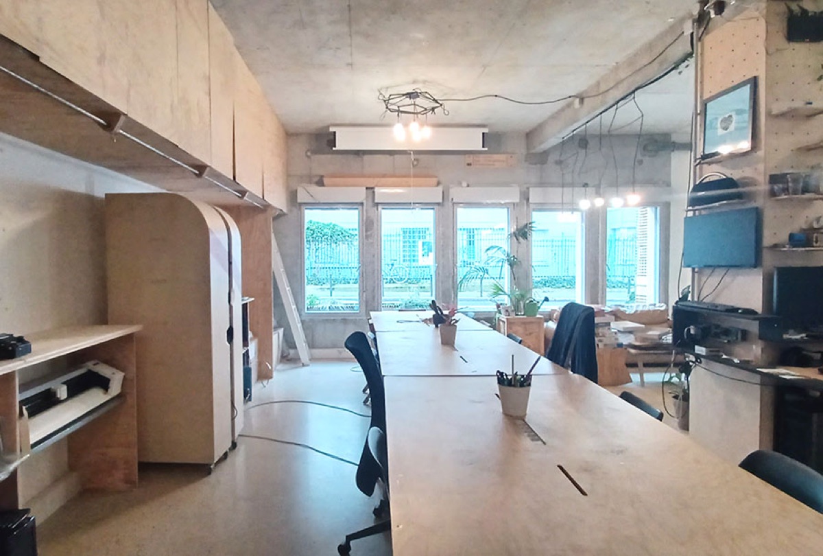 Espace de coworking nomade du WoMa : longue table en bois partagée avec chaises de bureau, murs en panneaux perforés garnis d’étagères, traceur sur un meuble d’atelier à gauche et grandes fenêtres donnant sur de la verdure